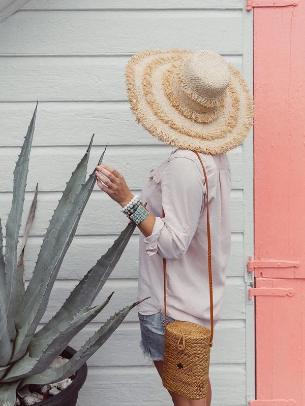 Woman in a large straw hat and light outfit next to a large succulent plant and pink door.