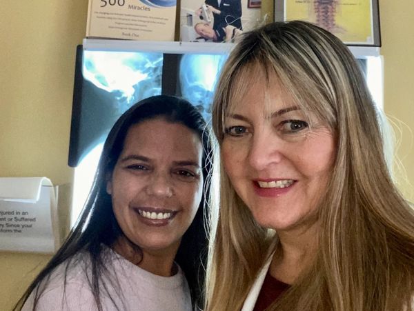 Two women smiling in a chiropractic clinic with spine X-rays in the background.