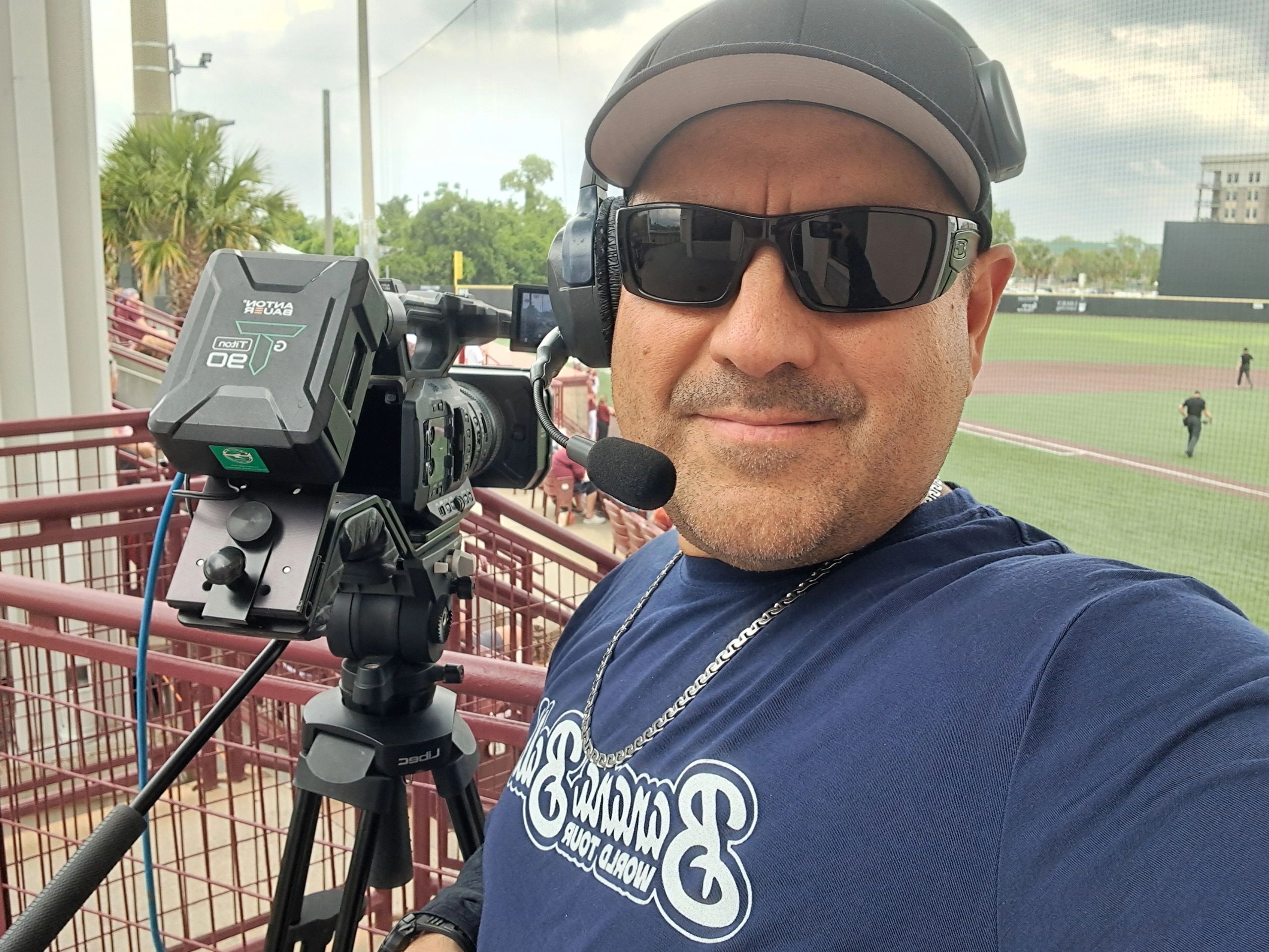 Man wearing sunglasses and a headset operates a video camera at a sports event.