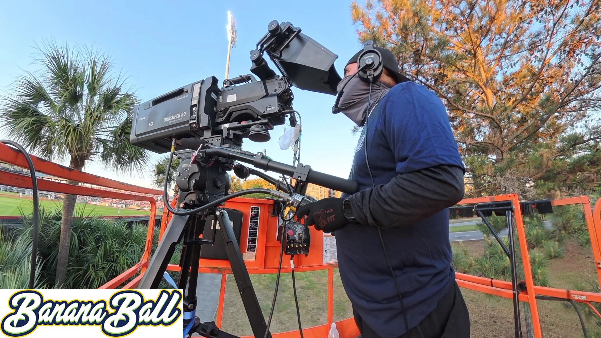 Camera operator filming an outdoor baseball game from a raised platform.
