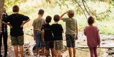 7 children standing in a line with their backs to the camera staring at a creek