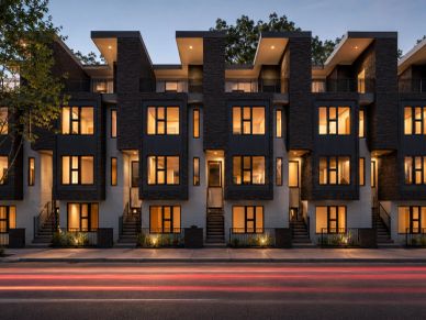 Modern four-story brick townhomes with illuminated windows along an urban street at dusk.
