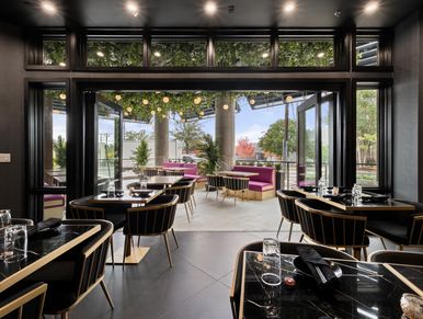 Modern restaurant interior with black marble tables, gold-trimmed chairs, and open glass doors to a 