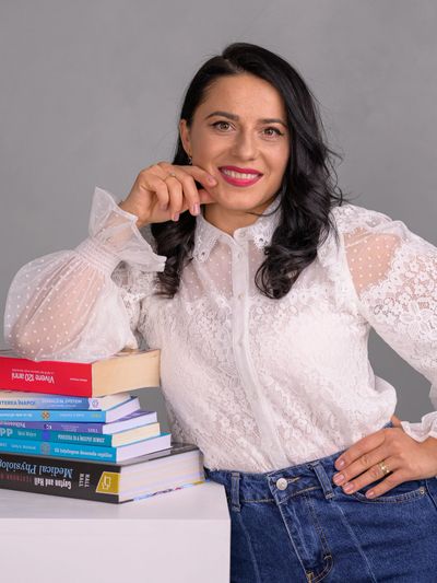 Confident woman in white lace blouse leaning on a stack of books, smiling.