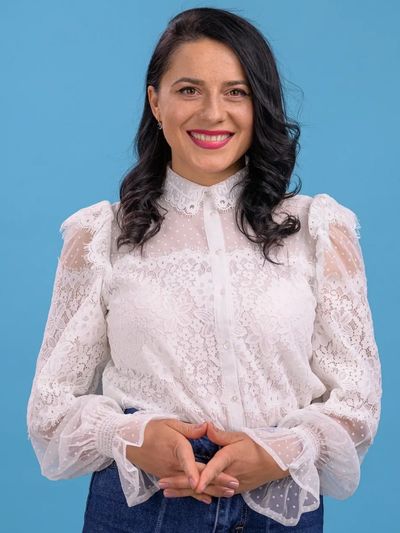 Woman smiling, wearing a white lace blouse against a blue background.
