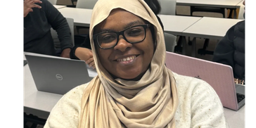 Smiling woman wearing glasses and a beige hijab in a classroom.