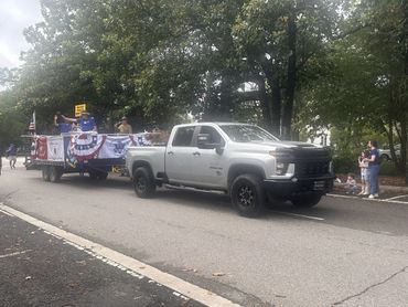 A silver pickup truck pulling a patriotic parade float on a tree-lined street.