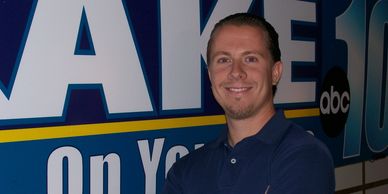 A man smiles standing in front of a news station sign.