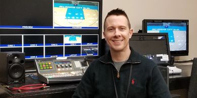 Man smiling in a broadcast control room with multiple monitors and equipment.