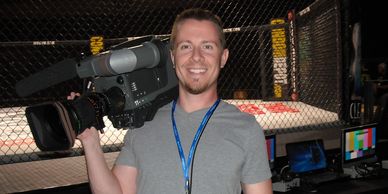 Man holding a professional video camera inside an MMA cage venue.