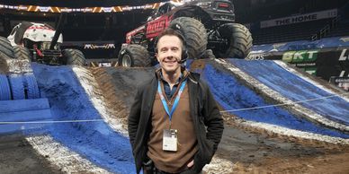 Man smiling at a monster truck event with trucks on a dirt ramp behind him.
