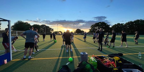Group of people playing field hockey during sunset on an outdoor pitch.