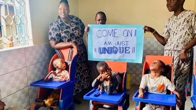Three children with special needs in high chairs, two adults holding a sign.