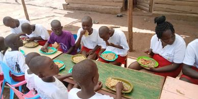 Children sitting and eating together at a wooden table outdoors.