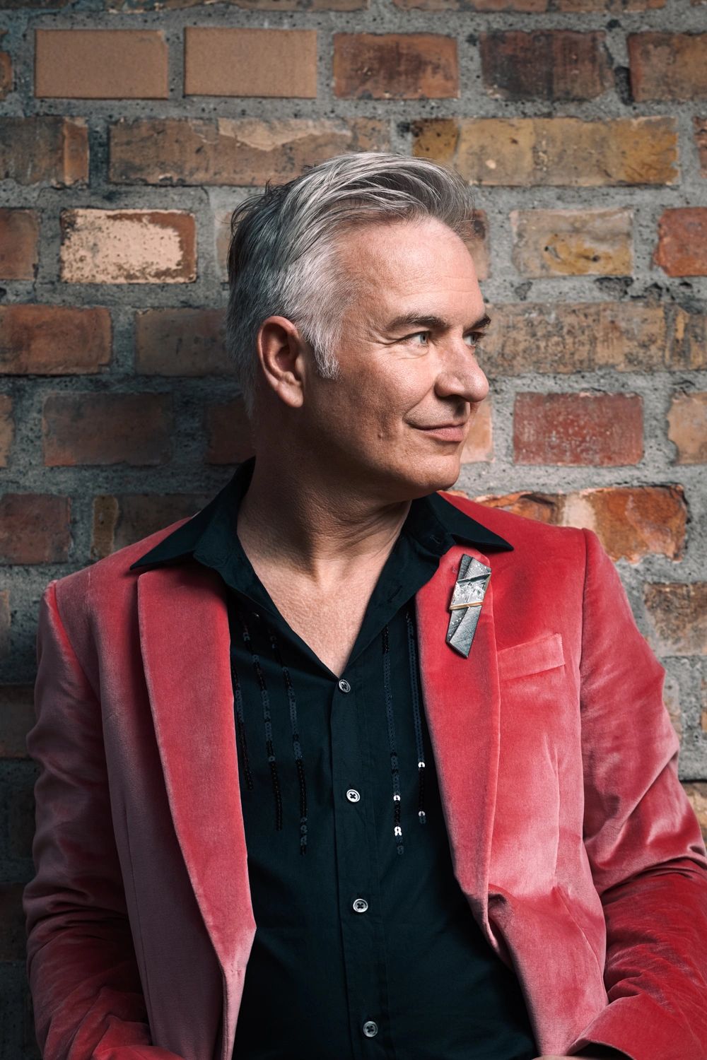 Man in a red velvet blazer with silver hair against a brick wall.