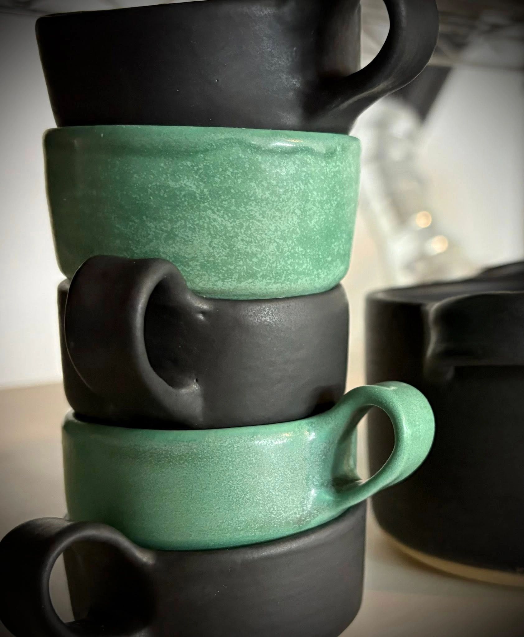 Tasses à anse faite à la main, de couleur vert océan empilées sur des tasses noir mat. 