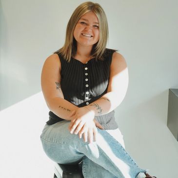 Smiling woman sitting on a stool in casual attire with natural sunlight.