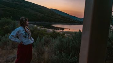 A woman gazes thoughtfully at a lake and mountains during sunset.