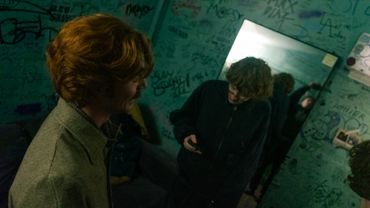 Two young men in a graffiti-covered room, one looking at a phone.