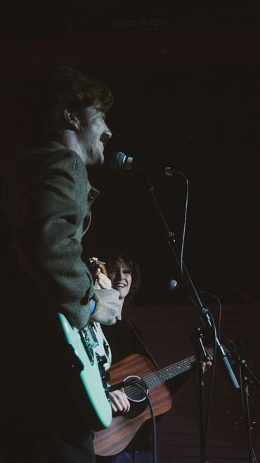 Two musicians play guitars and sing into microphones on stage.