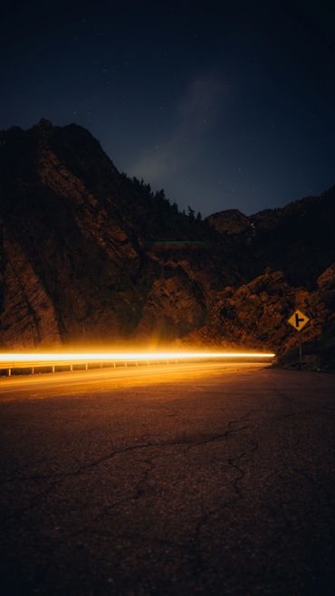 Long exposure of car lights on a mountain road at night under a starry sky.
