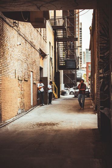 Three people in an urban alley with brick walls and fire escapes.