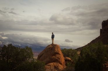 A person stands alone on a large red rock under a cloudy sky.