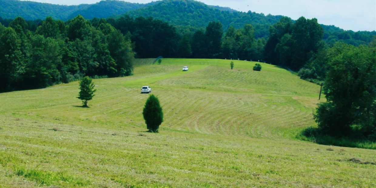 Two cars driving on a grassy field surrounded by trees and hills.
