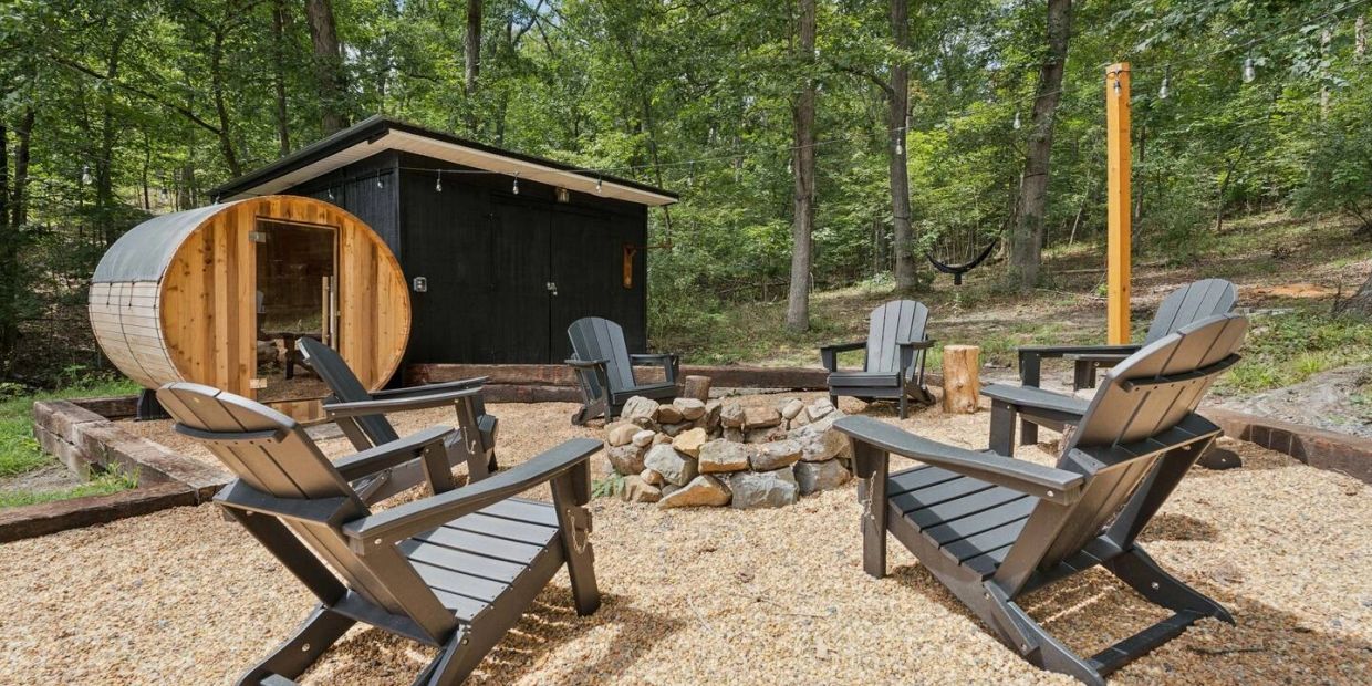 Outdoor seating area with wooden chairs around a stone fire pit near a barrel sauna in the woods.