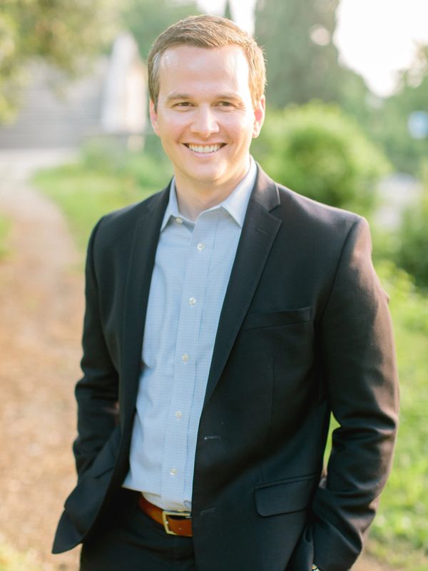 Smiling man in a black blazer and light blue shirt stands outdoors.