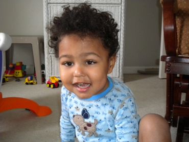 Pic 4.
Curly hair toddler in light blue pj's sitting on the floor and smiling for the camera.
