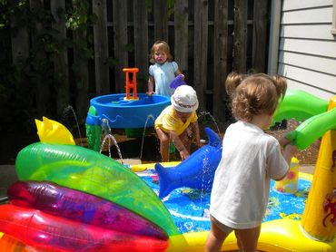 Pic 6.
3 Daycare kids playing in a backyard. One child plays at a water table, others 2 on water mat