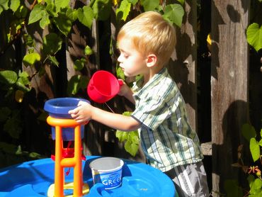 Pic 15.
Toddler stands at blue, orange and red water table holding a red cup in one hand.