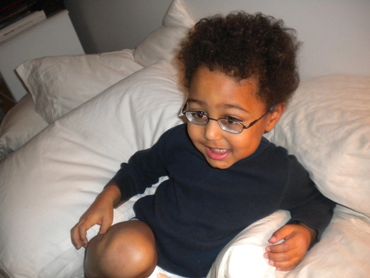 Pic 20.
Cute curly hair toddler with glasses sits among white bed pillows smiling happily