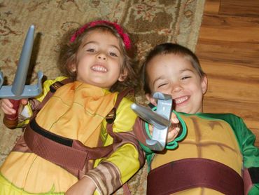 Pic 5. 
Girl and boy lying on the floor, wearing ninja turtle costumes and pointing a toy sword.