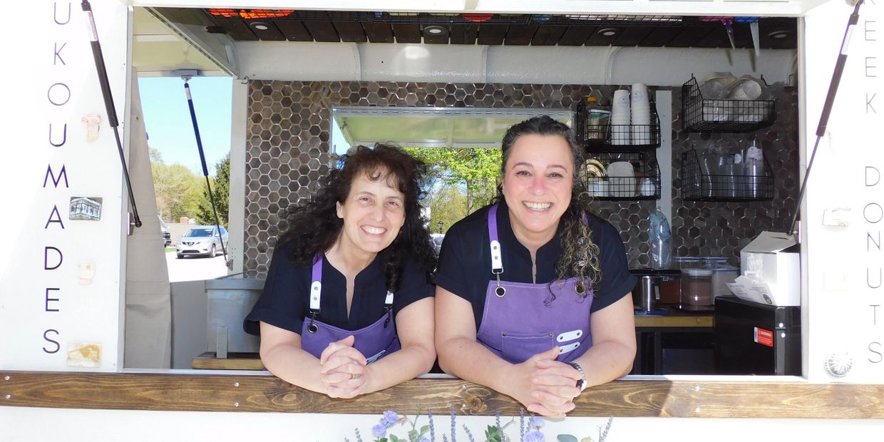 Two women smiling at a food truck window wearing purple aprons.