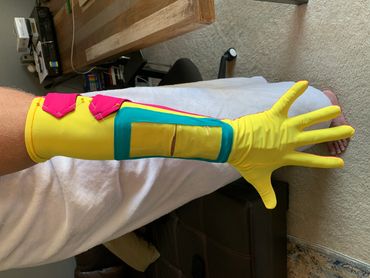 Bright yellow glove with pink and blue accents worn on arm indoors.