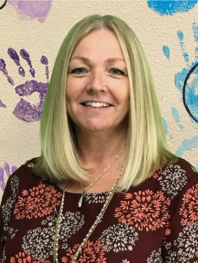 Smiling woman with blonde hair standing against a colorful handprint wall.
