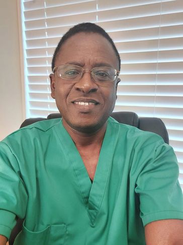 Man in green medical scrubs smiling indoors with blinds behind.