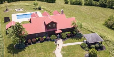A countryside house with a red roof, a pool, and parked cars in a green, wooded area.