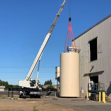 Crane lifting a large cylindrical tank next to an industrial building.