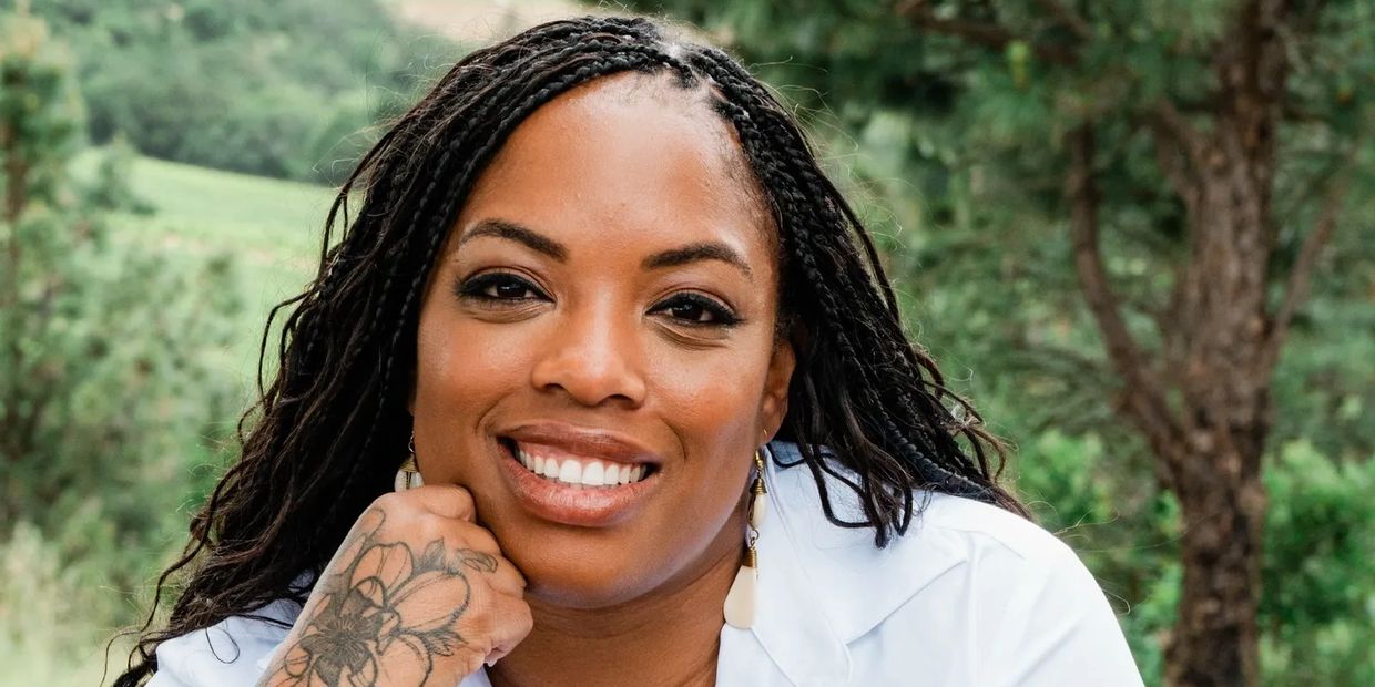 Smiling woman with braided hair and floral tattoo near wine bottles.