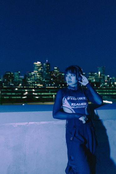 Woman in stylish outfit listens to music on headphones at night with city skyline behind.