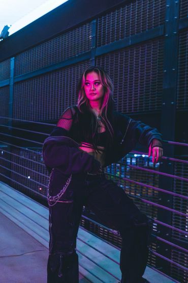 Young woman with neon lighting leans on railing in edgy outfit.