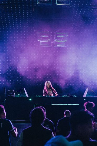 Female DJ performing on stage with purple lighting and crowd in front.