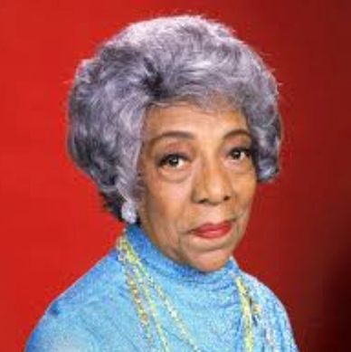 Elderly woman with gray hair and blue outfit against red background.