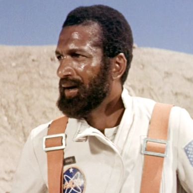 Man in a white astronaut suit with NASA patches in a desert setting.