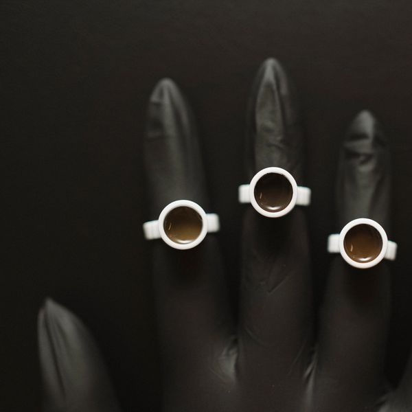 Black gloved hand with three espresso cups on fingertips.