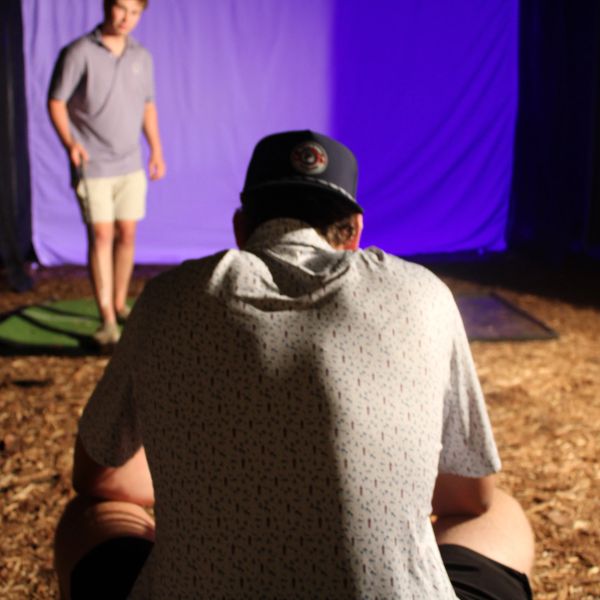 Two people, one sitting and one standing against a purple backdrop.