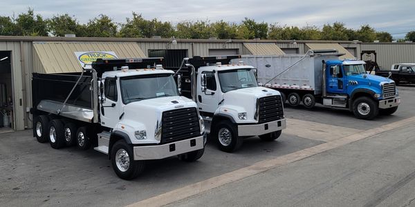 Three heavy-duty dump trucks parked outside a metal building under a cloudy sky.